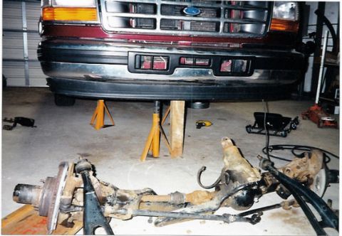 1996 Ford Bronco - The front end of a maroon Ford truck is elevated on jack stands, with its front axle removed and lying on the garage floor.