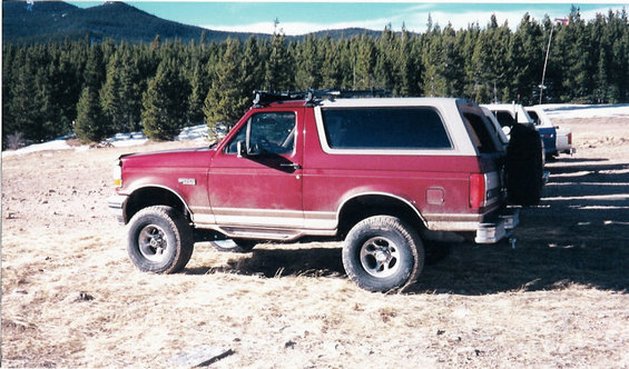 Red 1996 Ford Bronco parked in a dry, grassy area with trees and mountains in the background. Another vehicle is visible behind it
