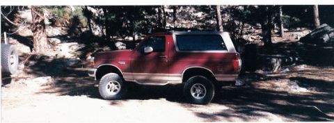 1996 Ford Bronco - A red Ford Bronco is parked outdoors on a dirt road, with trees and rocks in the background.