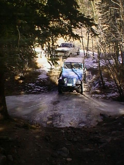 1996 Ford Bronco - Two off-road vehicles navigate a challenging dirt path through a wooded area.