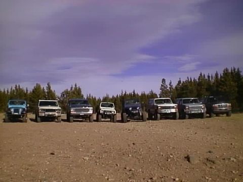 1996 Ford Bronco - A group of SUVs and trucks are parked in a line on a dirt lot with trees in the background.