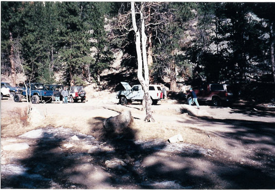 Several SUVs and off-road vehicles parked on a dirt lot in a forest. One vehicle has its hood open