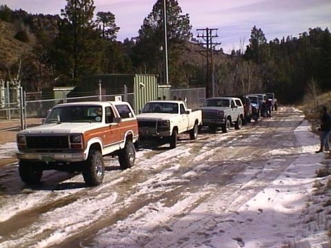 1996 Ford Bronco - A convoy of trucks and SUVs travels down a snowy, unpaved road near a utility substation.