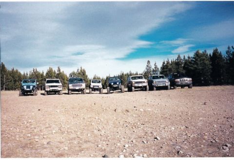 1996 Ford Bronco - A group of SUVs and trucks are parked in a line on a dirt lot, with trees and a blue sky behind them.