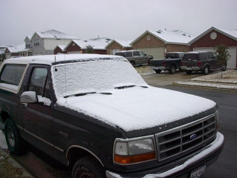 1992 Ford Bronco - Front three-quarter view of a dark green Ford Bronco covered in a layer of fresh snow on its hood and roof, parked at the curb of a suburba…