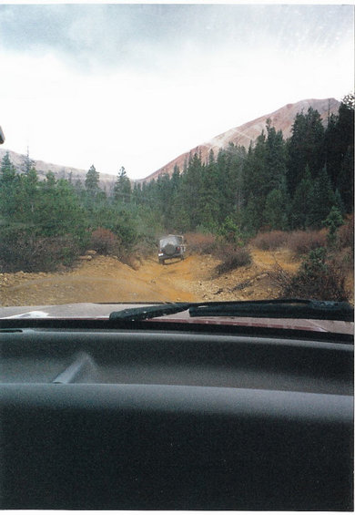 1996 Ford Bronco - Two off-road vehicles navigate a dusty trail through a wooded mountain landscape under an overcast sky.