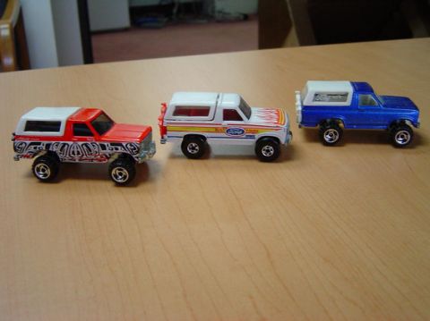 1996 Ford Bronco - Three die-cast toy 4x4s lined up in profile on a light wood tabletop: a red-and-white Bronco-style SUV with graphics, a white Ford Bronco w…