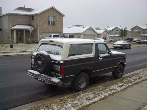 1992 Ford Bronco - Rear three-quarter view of a dark green Ford Bronco with a white removable hardtop, dusted with snow and parked at a suburban curb. A rear-…