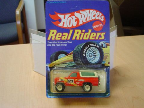 1996 Ford Bronco - Sealed Hot Wheels "Real Riders" blister card by Mattel on a wood table, holding a red-and-white Bronco-style 4x4 die-cast with a number 21…