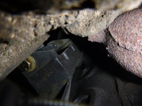 1953 Plymouth Belvedere - Underside view of a vehicle's engine component, showing a black bracket secured by a gold bolt amidst rusty exhaust pipes and grime.