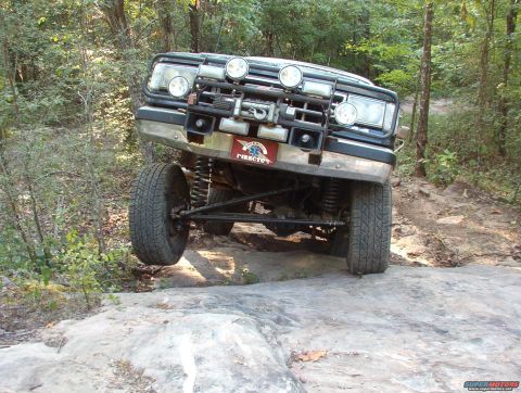1988 Ford Bronco - An off-road vehicle navigates a rocky trail with its front-right wheel lifted high.
