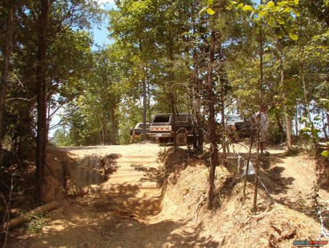 1988 Ford Bronco - Several off-road vehicles are parked on a dirt path surrounded by trees, with a challenging terrain ahead.
