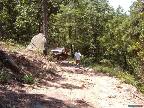 1988 Ford Bronco - A person walks along a rough, rocky path in a wooded area as an off-road vehicle navigates the terrain.