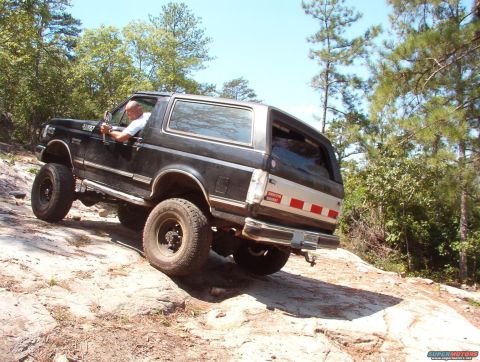 1988 Ford Bronco - A man drives a lifted Ford Bronco over rough terrain on a sunny day.