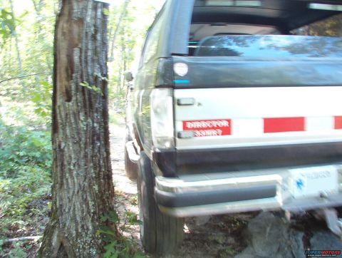 1988 Ford Bronco - Rear view of a black and white SUV, partially obscured by a tree, with a 'DIRECTOR' sticker visible on its side.