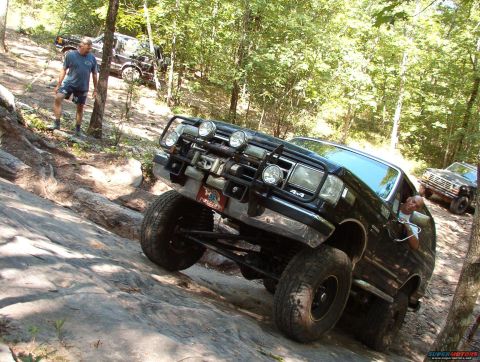 1988 Ford Bronco - Two Ford Broncos navigate a challenging rocky off-road course in a wooded area.