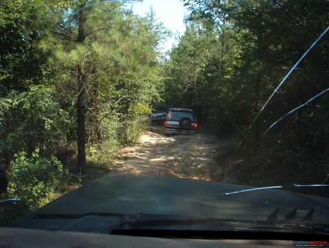 1988 Ford Bronco - A convoy of SUVs navigates a wooded trail, kicking up dust on the unpaved path.
