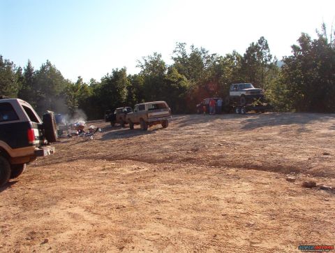 1988 Ford Bronco - Several off-road vehicles are gathered in a dirt lot with trees in the background.