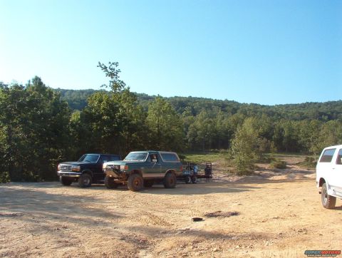 1988 Ford Bronco - Three Ford vehicles, including a green 1988 Bronco, are parked in a dirt lot with a wooded hill in the background.