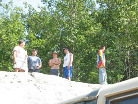 1988 Ford Bronco - A group of people stand on a sandy incline, with a vehicle's hood in the foreground and trees behind them.