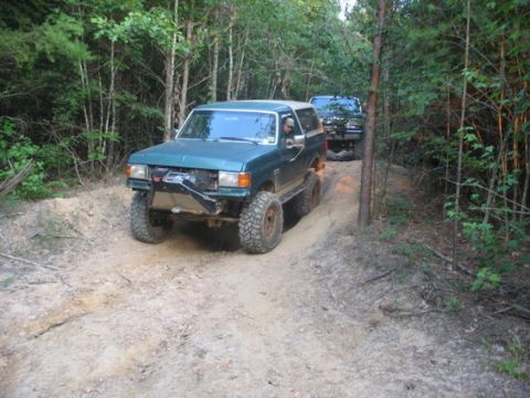 1988 Ford Bronco - A lifted, dark teal Ford Bronco is driving on a dirt trail through a wooded area. Another black SUV is following behind it.