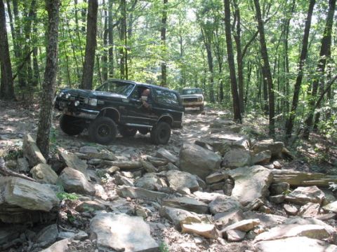 1988 Ford Bronco - A black Ford Bronco navigates a rocky, wooded trail, with a second vehicle visible further down the path. The Bronco is lifted and appears…