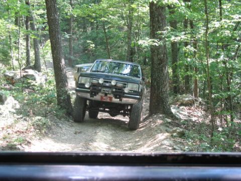 1988 Ford Bronco - A dark-colored Ford Bronco is driving on a dirt trail through a wooded area. The vehicle has a brush guard with multiple lights mounted on…