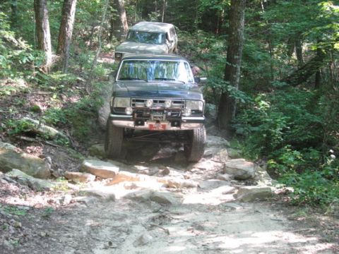 1988 Ford Bronco - A dark-colored Ford Bronco is navigating a rocky, wooded trail. Another light-colored SUV is visible behind it on the same path.