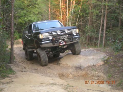 1988 Ford Bronco - A black, lifted 1980s Ford Bronco is navigating a rocky off-road trail with its front right tire on a large rock. The vehicle has a brush g…