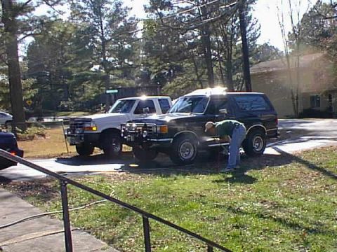 1988 Ford Bronco - A person in a green shirt and blue jeans is washing a black Ford Bronco parked next to a white Ford pickup truck on a sunny day.