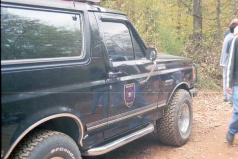 1988 Ford Bronco - A black Ford Bronco is parked on a dirt road with trees in the background. A search and rescue emblem is visible on the side door.