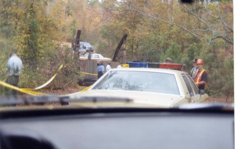 1988 Ford Bronco - A yellow police car with its light bar illuminated is parked behind a wooden structure in a wooded area, with yellow crime scene tape visib…