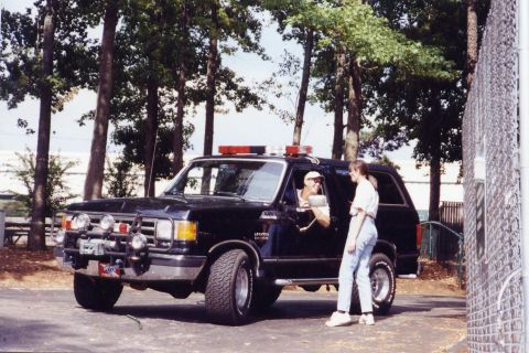 1988 Ford Bronco - A black Ford Bronco SUV with emergency lights on the roof is parked on a paved surface next to a chain-link fence. A man in a white hat is…