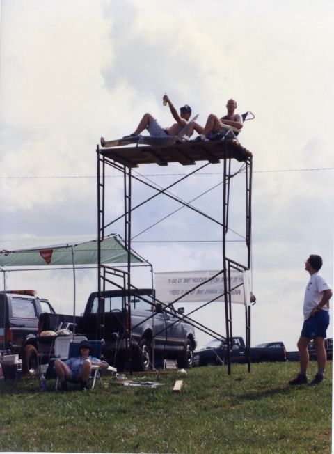 1988 Ford Bronco - Two men relax on a makeshift platform atop scaffolding, with one holding a bottle. In the background, several pickup trucks and an SUV are…