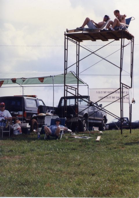 1988 Ford Bronco - Two men relax on a raised platform constructed from scaffolding, while two other people sit in chairs on the ground next to a black Ford Br…