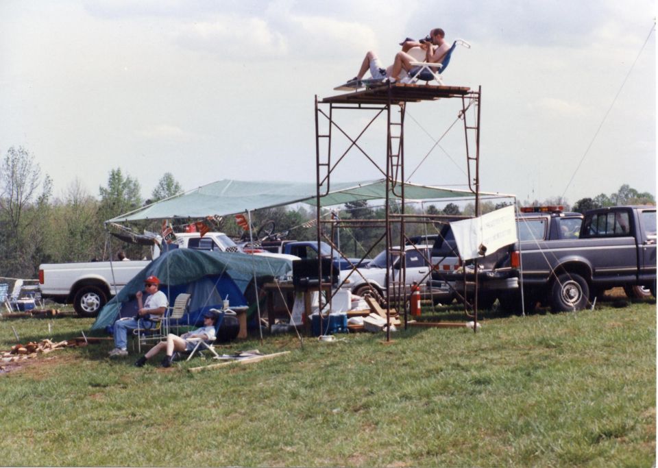 People at an outdoor event with tents and pickup trucks, two people on a raised platform.