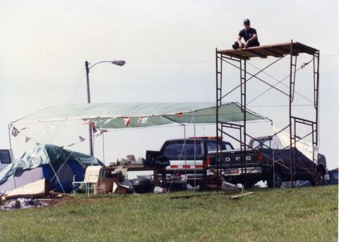 1988 Ford Bronco - A person sits atop a scaffolding structure next to a dark-colored Ford pickup truck parked at an outdoor event with tents and camping gear.
