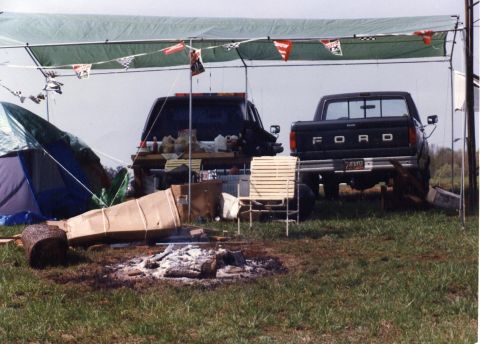 1988 Ford Bronco - A campsite scene with two dark-colored Ford pickup trucks parked under a green canopy. A tent is set up to the left, and a campfire pit is…