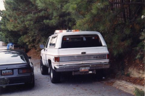 1988 Ford Bronco - The rear of a white Chevrolet SUV with a light bar on top is parked on a paved road next to a black car. The Chevrolet has a license plate…