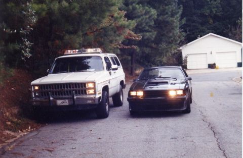 1988 Ford Bronco - A white SUV with emergency lights on the roof is parked next to a dark coupe on a paved road. A white two-car garage is visible in the back…