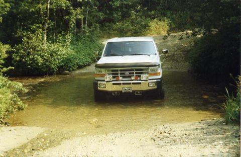 1988 Ford Bronco - A white Ford Bronco is driving through a shallow, muddy creek bed surrounded by dense green foliage. The front of the vehicle is facing the…