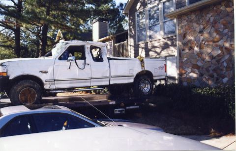 1988 Ford Bronco - A white Ford pickup truck is being towed on a flatbed trailer. The truck appears to have sustained significant damage to its roof and body.