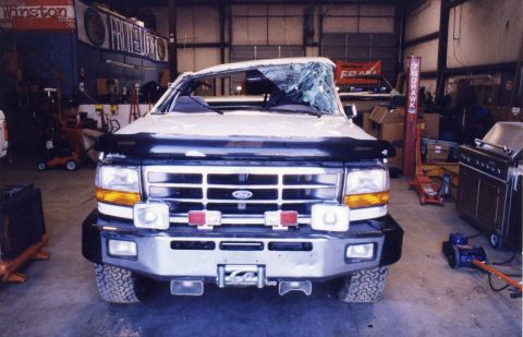 1988 Ford Bronco - A damaged white Ford truck with a shattered windshield is parked in a garage. The front of the truck features a chrome grille, aftermarket…