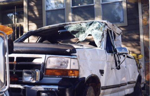 1988 Ford Bronco - A heavily damaged white Ford pickup truck with a shattered windshield and significant body damage is parked in front of a building with woo…