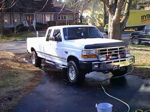 1988 Ford Bronco - A white Ford F-Series pickup truck is parked on a wet driveway in front of a house. A green hose is connected to the front bumper and runs…
