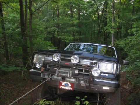 1988 Ford Bronco - A dark colored SUV is parked on a wooded trail, angled towards the camera. The front of the vehicle features a winch, multiple auxiliary li…