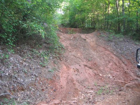1988 Ford Bronco - The front right corner of a black vehicle is visible on the right side of the image, parked on a dirt road that ascends a hill. The vehicle…