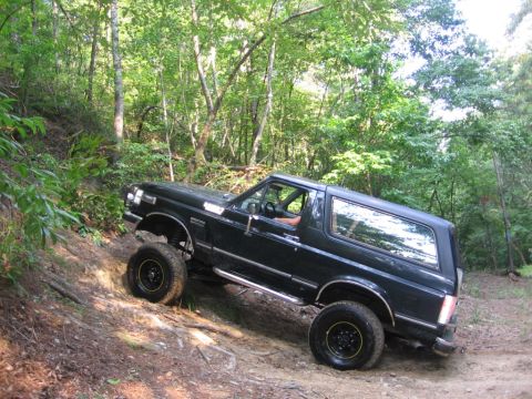 1988 Ford Bronco - A dark-colored Ford Bronco is shown from the side-rear angle, driving up a dirt trail in a wooded area. The vehicle is angled on the inclin…