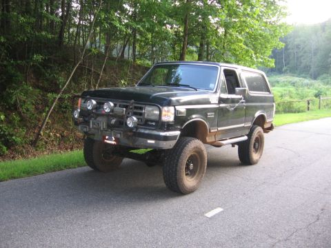 1988 Ford Bronco - A dark green, lifted 1988 Ford Bronco is parked on the side of a paved road, with a wooded area in the background. The front of the vehicle…