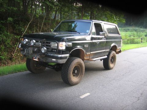1988 Ford Bronco - A dark green, lifted 1988 Ford Bronco is parked on an asphalt road next to a wooded area. The vehicle features a brush guard with multiple…